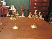 Pair of five-prong brass candelabras on a wooden table with chairs and furniture in background