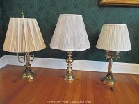 Three brass table lamps with pleated white shades arranged on wooden floor against a green patterned wallpapered wall.