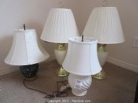 Photo showing all 4 decorative table lamps with varied bases and white lampshades, including matching pair and 2 misc lamps on carpeted floor near wall