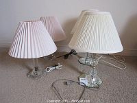 Four clear glass table lamps with faceted bodies and mirrored hexagonal bases, with pleated fabric lamp shades in white and off-white colors, cords visible on the carpeted floor.