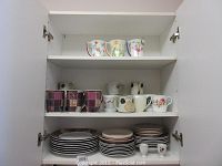 Full cupboard shot showing three shelves with assorted mugs, many white plates stacked below, and two rooster-illustrated egg cups on bottom shelf.