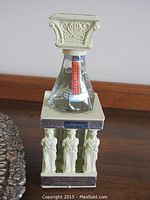 Greece-themed small wide bottle shaped with classical pillar topped by square capital, featuring sculpted female figures and painted details.