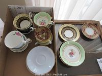 12 china cups and saucers arranged in box and glass display showing various patterns and colors including green, dark blue, and pink with gold accents.