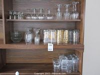 Wooden cabinet shelves holding various glassware items including wine glasses, tumblers with gold designs, and clear glass mugs and bowls.