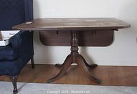 Drop leaf wood table shown with both leaves down, folded against the wall, pedestal base with four curved legs and caster wheels visible.