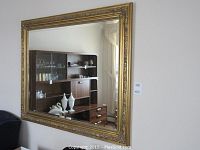 Large rectangular wall mirror mounted on wall with a gold ornate frame, reflects room interior.