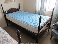 Antique wood single bed with mattress showing side and corner view, displaying turned finials and caster wheels.