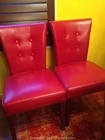 Two red tufted bonded leather chairs side by side against a yellow wall, showing upholstery and button details, wooden legs visible