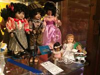 Photo of six dolls displayed on a shelf with various outfits: three larger dolls with dark hair and three smaller dolls including a blonde and a German folk outfit.