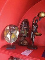 Three items displayed on shelf: two wooden masks and one bronze-colored figurine.