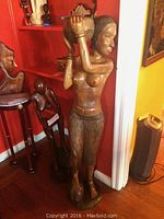 Full view of the carved wooden statue of an African woman carrying a bowl on her head, standing 50 inches tall, positioned in a room with red walls.