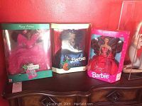 Three boxed Barbie dolls displayed side by side on a wooden surface against a red wall. From left to right: Holiday Barbie in pink with wings, Velvet and Jewels Barbie in black gown, Radiant in Red Barbie in red gown.