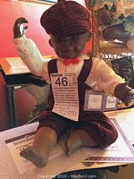 Full view of porcelain doll 'David' seated wearing red plaid vest, cap, white shirt, and red bow tie, with associated documentation underneath