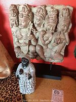 Photo showing the Hindu stone relief sculpture with five figures and the African carved figurine in front on a patterned surface against a red wall.