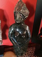 Front full view of the dark green jade female statue with arms crossed and textured headdress, displayed against a red background.