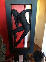 Full view showing the metal sculpture of a female figure posed inside a rectangular black frame, approximately 27 inches tall.