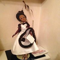 Full view of the Madame Alexander doll wearing the ivory satin and cafe rose colored cocktail dress with accessories, standing upright on stand.