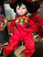 Full body view of Brinn's porcelain Asian doll dressed in a red shiny outfit with gold decorative details, seated on white display stand.