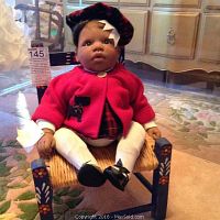 Full front view of the vinyl doll seated on the wicker chair showing outfit and face