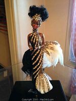 Full frontal view of Barbie doll on black stand inside clear plexiglass case showing gown with black and gold beadwork and feathered train.