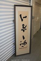 Full view of original Asian calligraphy scroll framed vertically, showing bold black brush strokes and artist signature at bottom.