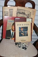 Photo showing two hardcover books, 'Four Days' and another JFK book, a collection of newspaper clippings bundled by string, and some small trading cards placed on a chair.