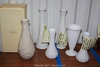 Photo showing the full assortment of one Lenox vase with embossed design and gold rim, and five milk glass vases with varying textures and heights, all arranged on a wooden surface.