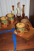 Four coffee mugs and two square plates with sunflower and brown swirl pattern, along with four blown glass bud vases in amber and purple hues on wooden surface.