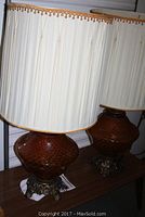 Pair of large amber glass lamps with cream pleated shades with fringe, on dark ornate metal bases on a wooden surface.