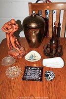 Photo showing wooden decanter with six small wooden cups and wooden tray, gold toned vase about 13 inches tall, kissing couple statue with some paint loss, and six assorted ashtrays of various materials and styles.