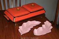 Orange mid century chafing dish with brass legs and covers shown with pink speckled leaf-shaped serving dishes on wooden table.