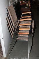 Four brown tweed fabric cushioned stackable chairs with metal frames and wooden armrests, stacked against a wall.