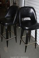 Two black mid-century modern vinyl covered swivel bar stools with metal legs shown side by side, visible wear, stains, and holes in vinyl upholstery.