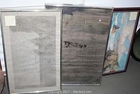 Three framed artworks leaning against wall: two silver-framed drawings and one wood-framed seascape.