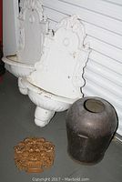 Two white plaster wall fountains with ornate backs and basins, showing paint chipping, placed on the floor next to a large dented dark metal urn and a gold-colored carved fruit bowl plaque