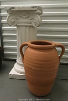 Large terra cotta planter pot in reddish-brown color with two handles, wide opening, and drainage hole visible at bottom.