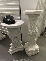 Two white plaster pedestals side by side, one with floral motif, one with angels, both showing some paint chips. Small black wall planter is on the taller pedestal.
