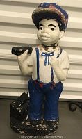 Front view of the painted cement statue of a boy holding a shoe and shoe shine brush, showing significant paint chipping on the cap, pants, and base.