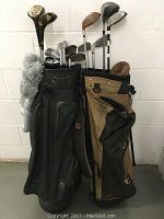 Two golf bags side by side filled with golf clubs including woods, irons, and putter, against a white wall.