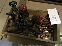 Photo of multiple copper and brass valves with colored handles inside a wooden box, showing gate valves and ball valves.