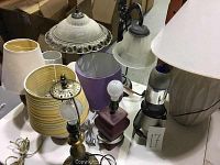 Photo of nine lamps with assorted shades including fabric and metal shades, with some damaged or missing shades visible.