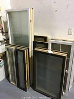 Photo showing various fixed single pane windows, some with frosted glass, in black and beige aluminum-clad frames stacked against a wall.