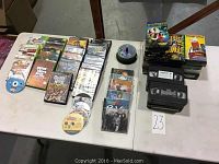 Photo showing a collection of video games, CDs, and VHS tapes arranged on a table with a label numbered 23. Includes PlayStation 2 game cases, DIY music CDs and stack of VHS tapes.