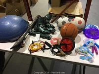 Full view of sports items on white table showing half exercise ball, inline skates, skateboard, balls, skipping ropes, frisbees, and ab roller wheel