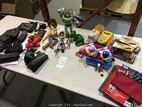 Full table view showing pull string Woody, Buzz Lightyear figure, green dinosaur, small action figures, toy cars, slot car racetrack sections, craft supplies including coloring books, colored pencils, markers, glue sticks, spiral notebooks.