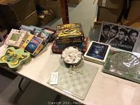 Photo of the overall lot showing board games stacked, a glass chess set on a glass chessboard with frosted and clear pieces, and the signed band posters behind.
