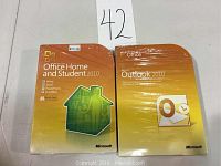 Two boxed Microsoft software packages: Office Home and Student 2010 and Outlook 2010, both shrink-wrapped with price tag visible on Office Home and Student 2010 box