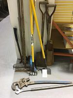 Full view of garden tools including axes, rakes, shovels, hoe, and broom leaning against a wall with pipe wrenches and a wrench on the floor.