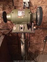 Front view of the entire bench grinder mounted on a pedestal, showing the grinding and brush wheels, on/off switch, and pedestal with some paint wear.