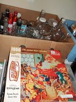Top view of box containing several clear and metallic Coca-Cola bottles along with a Coca-Cola 1000 piece jigsaw puzzle box.